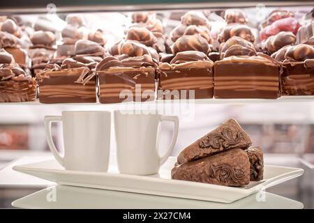 Drei Stücke Schokoladenwaffelkuchen mit zwei Tassen auf einer weißen Keramikplatte, auf dem Hintergrund einer Vitrine mit Süßigkeiten. Stockfoto