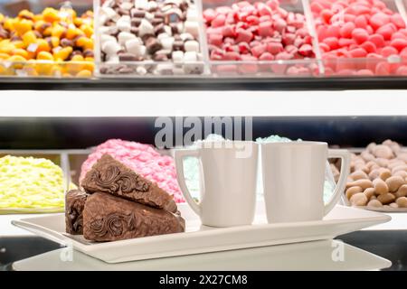 Drei Stücke Schokoladenwaffelkuchen mit zwei Tassen auf einer weißen Keramikplatte, auf dem Hintergrund einer Vitrine mit Süßigkeiten. Stockfoto