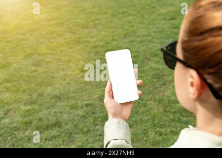 Frau, die ein Smartphone mit einem weißen Bildschirm in der Hand vor grünem Rasen hält. Mockup-Bildschirm des Telefons über der Schulter. Stockfoto