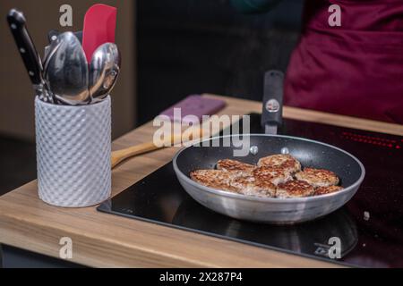 Knusprige Fleischbällchen in einer Pfanne auf einem modernen Induktionsherd, Küchenutensilien in einem Halter an der Seite, was auf ein aktives kulinarisches Ambiente hindeutet. Hoch qua Stockfoto