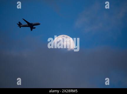 Ein kommerzielles Flugzeug, das sich auf einem Flughafen befindet, überquert einen Vollmond, der in der Abenddämmerung teilweise von niedrigen Wolken verdeckt ist. Stockfoto