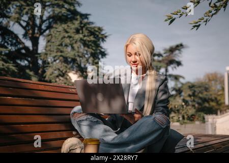 Eine blonde Frau sitzt auf einer Bank mit einem Laptop vor sich. Sie trägt eine graue Jacke und Jeans. Die Szene deutet auf eine lässige und entspannte Atmosphäre hin Stockfoto