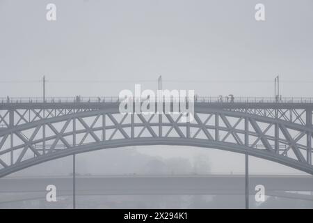 Menschen mit Regenschirmen und Regenmänteln, die auf dem oberen Bahnsteig der Don Luis I Stahlbrücke laufen, und Autos, die auf dem unteren Bahnsteig in Porto fahren. Stockfoto
