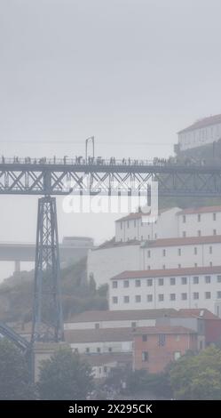 Touristen und Fußgänger, die mit Regenschirmen und Regenmänteln auf der oberen Plattform der Dom-Luis-Brücke wandern, umgeben von Nebel und niedrigen Regenwolken. Stockfoto