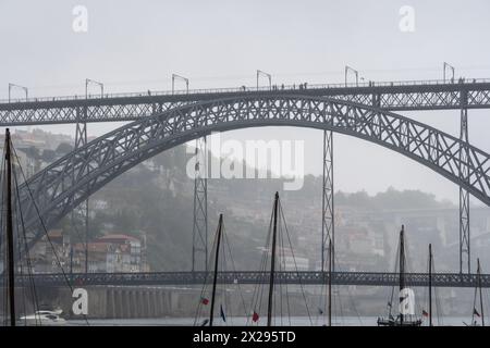 Masten von hölzernen Ragelos-Booten, die auf dem Fluss Douro in Porto unter der Stahlbrücke Don Luis I im Hintergrund angedockt sind, umhüllt von Nebel bei grauem Regen Stockfoto