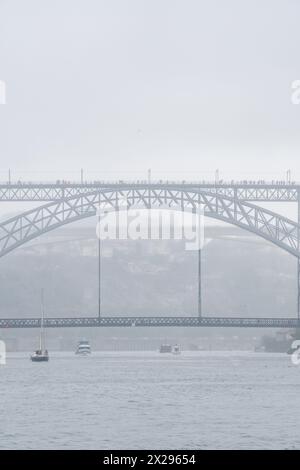 Die Stahlbrücke Don Luis I ist in Nebel gehüllt und voller Touristen, die mit Regenschirmen und Regenmänteln an einem sehr nebeligen und regnerischen Tag mit Booten unterwegs sind Stockfoto