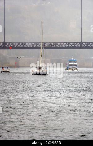 Die Touristenfähre und ein Boot, das entlang des Douro-Flusses fährt und an einem sehr nebeligen und regnerischen Tag unter der Don Luis I-Brücke in Porto vorbeifährt. Stockfoto