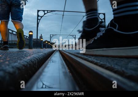 Nahaufnahme der Herrenfüße mit Turnschuhen und Shorts, die bei Sonnenuntergang auf der Dom Luis-Brücke die Metro von Porto überqueren, mit der Reflexion der Kabel und Stockfoto