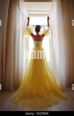 Die Silhouette einer Frau in einem goldenen, luxuriösen Kleid vor dem Hintergrund eines Fensters hält einen Vorhang mit den Händen. Elegante Dame in gelber Seide Stockfoto