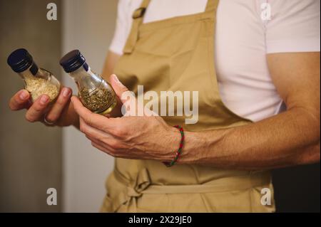 Die Hände des Küchenchefs halten zwei Glasflaschen mit Gewürzen, getrockneten Basilikumblättern und einem Gewürzgericht während des Abendessens Stockfoto