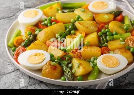 Salat mit Bratkartoffeln, Spargel, gekochtem Ei, Tomaten und Erbsen auf dem Teller auf dem Tisch. Horizontal Stockfoto