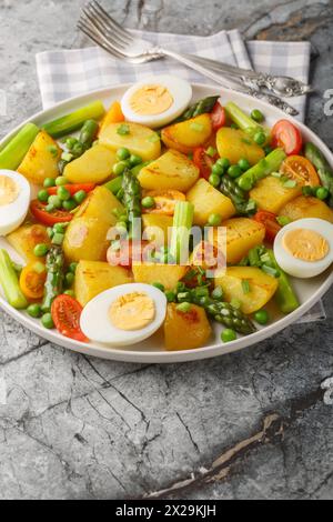 Sommersalat mit Kartoffeln, grünen Erbsen, Spargel, gekochtem Ei und Tomaten auf dem Teller auf dem Tisch. Vertikal Stockfoto