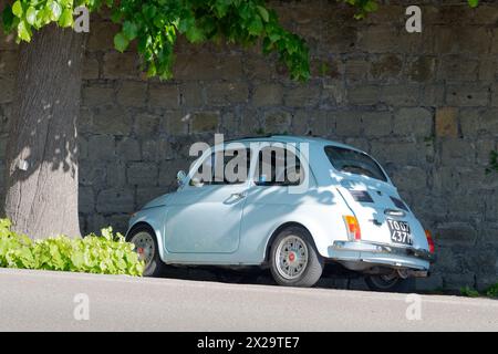 Hellblaues Classic Fiat 500, das neben einem Baum und einer hohen Steinmauer im Schatten in Montefiascone, Italien, geparkt wird. April 2024 Stockfoto