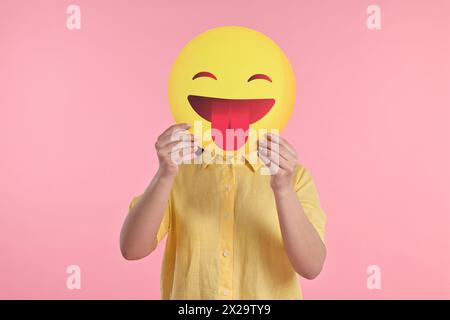 Frau, die das Gesicht mit Emoticon bedeckt und die Zunge auf rosa Hintergrund hinausragt Stockfoto