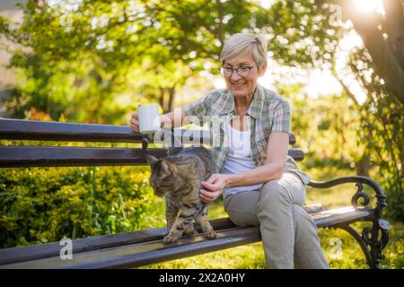 Glückliche Seniorin trinkt gern Kaffee und verbringt Zeit mit ihrer Katze, während sie auf einer Bank in ihrem Garten sitzt. Stockfoto