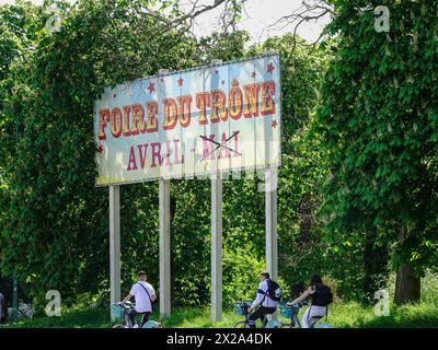 Jugendliche auf dem Fahrrad passieren das Foire du Trône-Schild, das den Wechsel vom regulären Monat Mai zu einem früheren April aufgrund der Olympischen Spiele ankündigt. Stockfoto