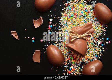 Schokoladeneier und Kaninchen, zuckerfarbene Streusel auf schwarzem Hintergrund, Draufsicht. Osterkomposition. Osterschokolade-Häschen. Festliche süße Köstlichkeiten. C Stockfoto