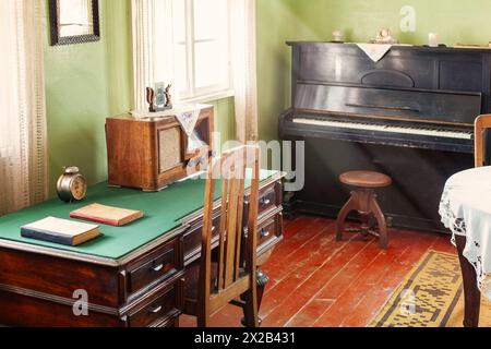 Antikes Arbeitszimmer mit Schreibtisch, Büchern, Uhr, Radio und schwarzem Klavier. Der Innenraum wird durch Tageslicht beleuchtet. Stockfoto