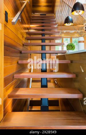 Blick auf eine beleuchtete Holztreppe mit geöffneten Stufen und durchsichtigem Glasgeländer, die zum oberen Stockwerk in luxuriösem gebeiztem Zedernholz und Holz führt Stockfoto