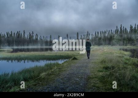 Wanderung am Vormittag am nebeligen Teich in der norwegischen Wildnis. Rückansicht der Frau macht einen friedlichen Spaziergang entlang eines Weges an einem nebelumhüllten Waldsee in Norwegen. Stockfoto