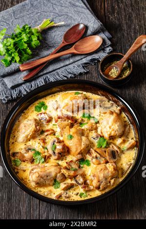 Hähnchenschenkel in einer cremigen Pilz-Knoblauchsauce mit Kräutern und Parmesan in Backform auf dunklem Holztisch mit Holzlöffeln, vertikale Ansicht Stockfoto