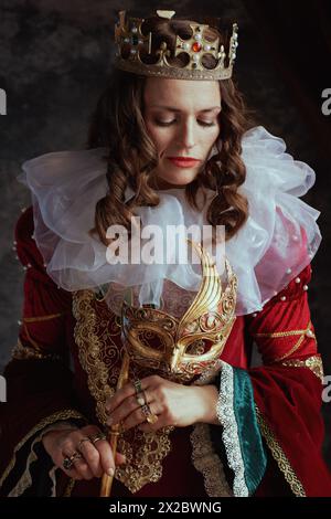 Nachdenkliche mittelalterliche Königin in rotem Kleid mit venezianischer Maske, weißem Kragen und Krone auf dunkelgrauem Hintergrund. Stockfoto