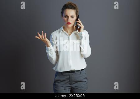 Gestresste, stilvolle 40-jährige Arbeiterin in weißer Bluse mit einem Handy isoliert auf Grau. Stockfoto