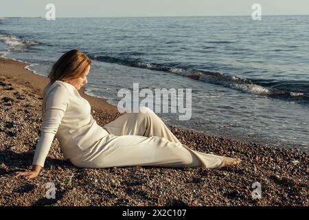 Eine Frau sitzt an einem Sandstrand neben dem Meer und blickt auf die Wellen, die unter einem klaren blauen Himmel gegen die Küste krachen. Stockfoto