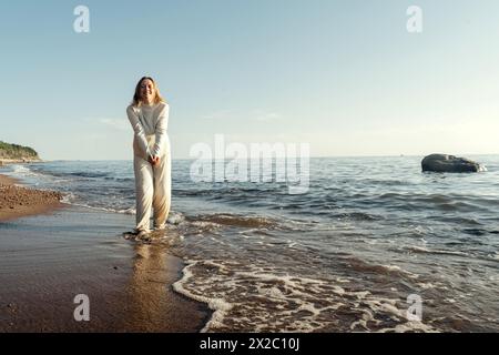 Eine Frau steht im Wasser am Strand, mit Wellen, die sanft um sie herum laufen. Sie blickt zum Horizont unter dem klaren blauen Himmel. Stockfoto