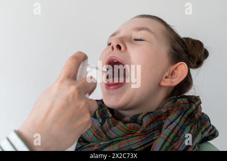 Ein kleines Mädchen wird mit Medikamenten in den Mund gesprüht. Ein Mädchen mit Halsschmerzen in einem Schal mit offenem Mund. Ausgewählter Fokus. Hochwertige Fotos Stockfoto