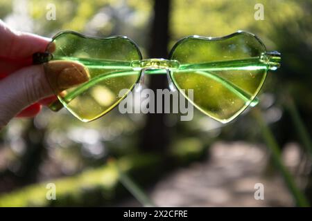 Eine Frau, die an sonnigen Sommertagen durch eine grüne, herzförmige Sonnenbrille auf die Natur schaut. Urlaub am Meer im tropischen Resort. Exotische Landschaft. Stockfoto