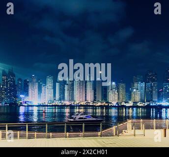 Abendlicher Meerblick von der Dubai Bluewaters Promenade zur Jumeira Beach Residence, Boot schwimmt auf Wasser und Mond unter Wolkenkratzern Stockfoto