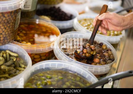 Die Hände der Herren halten die Kelle und legen eingelegte schwarze Oliven mit Stein in einen Einwegbehälter aus Kunststoff Stockfoto