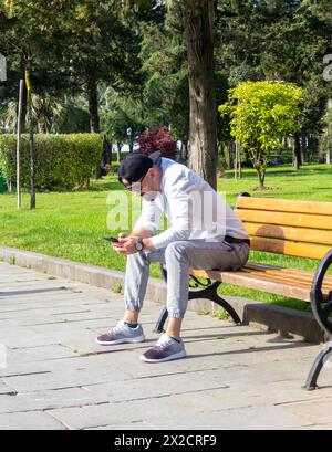 Batumi, Georgien. 04.16.2024 Ein Mann mit Mütze schaut auf einer Bank auf das Telefon. Entspannung im Park. Kommunikation in sozialen Netzwerken Stockfoto