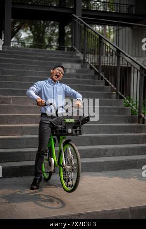 Ein überschwänglicher asiatischer Geschäftsmann fährt freudig mit einem grünen Fahrrad die Treppe hinunter und drückt Glück und Freiheit aus. Das Konzept der Geschäftsleute Stockfoto
