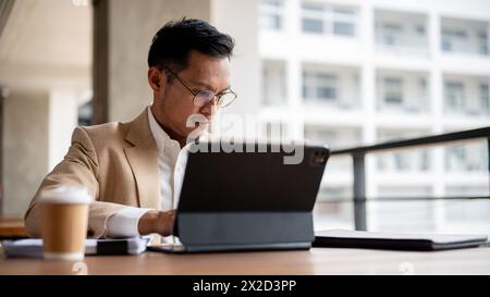 Ein zielgerichteter asiatischer Millennial-Geschäftsmann in beigefarbenem Anzug und Brille arbeitet an seinem digitalen Tablet und liest Geschäftsdokumente an einem Tisch in einem Stockfoto