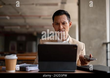 Ein verwirrter, ernsthaft aussehender asiatischer Geschäftsmann der Millennials starrt mit einem verwirrten Ausdruck auf seinen digitalen Tablet-Bildschirm und sieht sich während des Arbeitsgangs einem Problem gegenüber Stockfoto