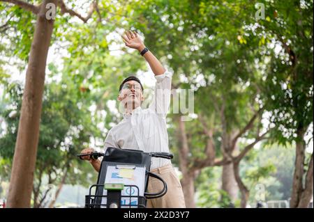 Ein begeisterter, glücklicher asiatischer Geschäftsmann der Millennials lächelt und winkt, während er sein Fahrrad in einem grünen Park schiebt und jemanden begrüßt, während er zur Arbeit aufbricht Stockfoto