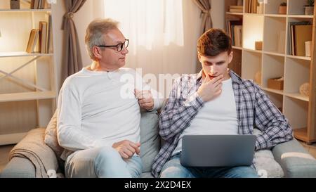 Elternunterricht. Familienhilfe. Der ältere Vater hilft dem verwirrten Sohn, das Arbeitsprogramm online auf einem Laptop-hellen Innenraum zu nutzen. Stockfoto
