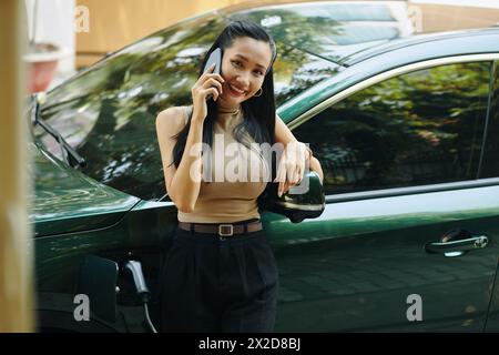 Lächelnde erfolgreiche Geschäftsfrau, die neben dem Elektroauto steht, wenn sie telefoniert Stockfoto