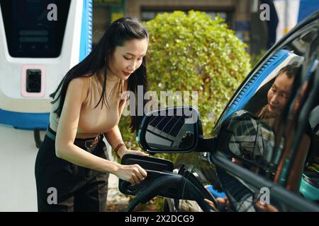 Eine Frau, die den Stecker in ihre EV steckt, wenn sie das Auto an einem öffentlichen Punkt auflädt Stockfoto