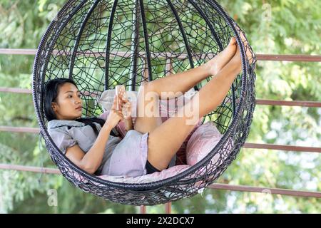 Eine junge Frau entspannt sich mit einem Handy in einem Hängesessel Stockfoto