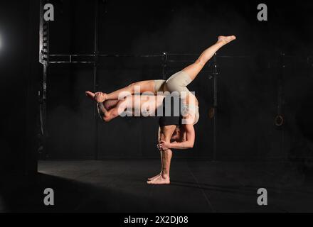 Acroyoga als Partnersportaktivität für junge Männer und Frauen. Ein Paar, das zusammen im Fitnessstudio trainiert und komplizierte Manöver durchführt. Stockfoto