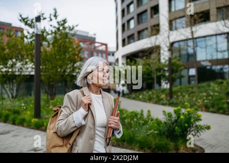 Reife Geschäftsfrau, die auf dem Smartphone scrollt und von der Arbeit nach Hause geht. Schöne ältere Frau mit grauen Haaren, die die Stadtstraße entlang läuft. Stockfoto