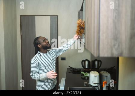 Ein lächelnder afrikanischer Mann wischt mit einem Federstaub Staub von den Möbeln ab. Konzept der allgemeinen Ordnung und Sauberkeit Stockfoto