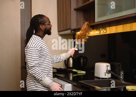 Junger afrikanischer Mann, der mit einem Federstaub Staub auf dem Schrank abwischt. Konzept der allgemeinen Ordnung und Sauberkeit Stockfoto