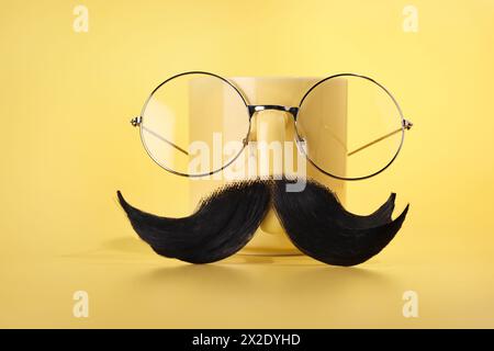 Männergesicht aus künstlichem Schnurrbart, Brille und Tasse auf gelbem Hintergrund Stockfoto