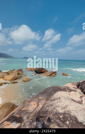 Klares Wasser prallt sanft gegen einen felsigen Strand unter einem hellblauen Himmel mit flauschigen Wolken Stockfoto