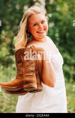 Strahlendes Teenager-Mädchen in weißem Sommerkleid, das Cowgirl-Stiefel hält Stockfoto