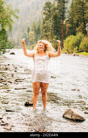 Fröhliches Teenager-Mädchen in weißem Sommerkleid, das vor Freude im Fluss springt Stockfoto